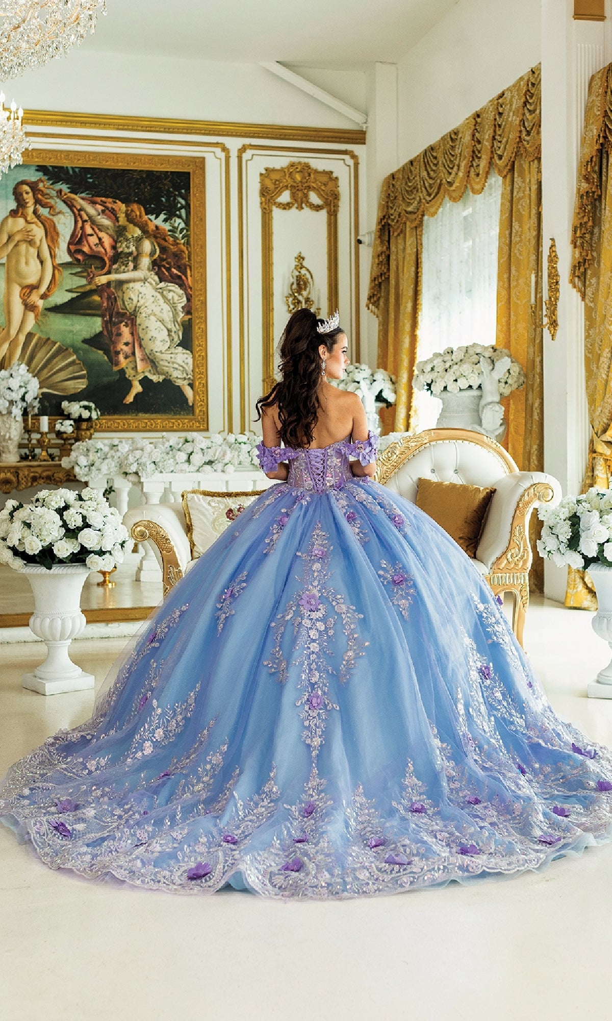 A woman wearing the Blue Quinceanera Dress 1999 By Dancing Queen, featuring a ball gown skirt and floral details, stands in an ornate room with gold décor and white flowers, facing away from the camera.